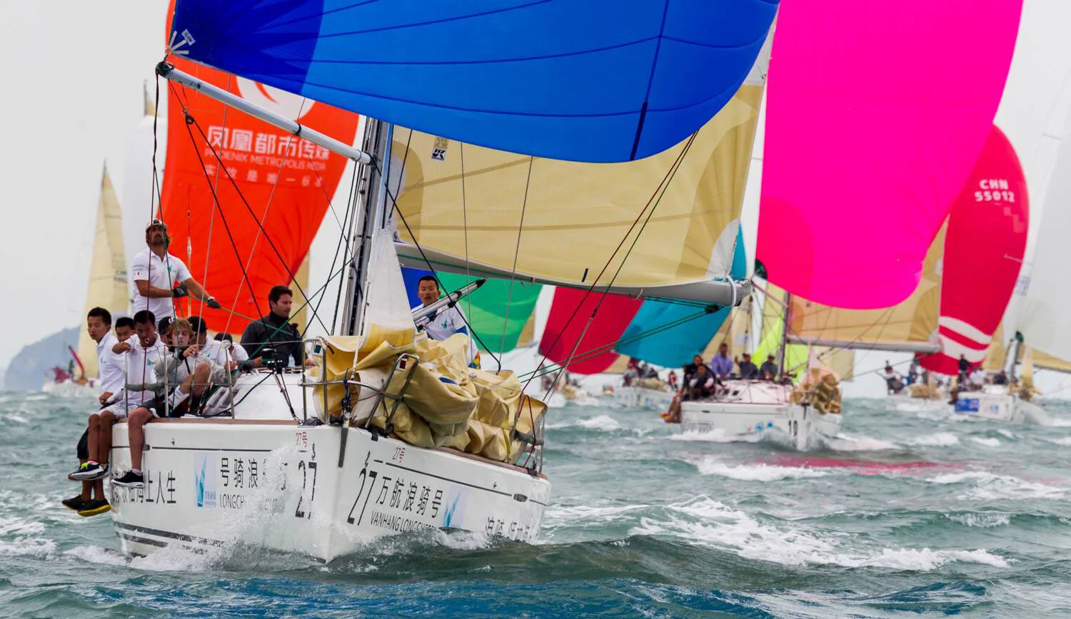 Racing fleet running under colourful spinnakers, crews on the bow of boat 27 with Chinese lettering on its hull