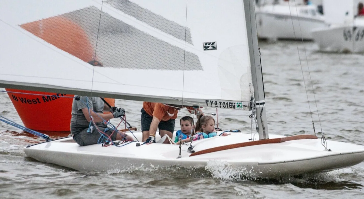 Two young children in the cockpit of a small keelboat with adults aboard, UK Sailmakers logo on the mainsail
