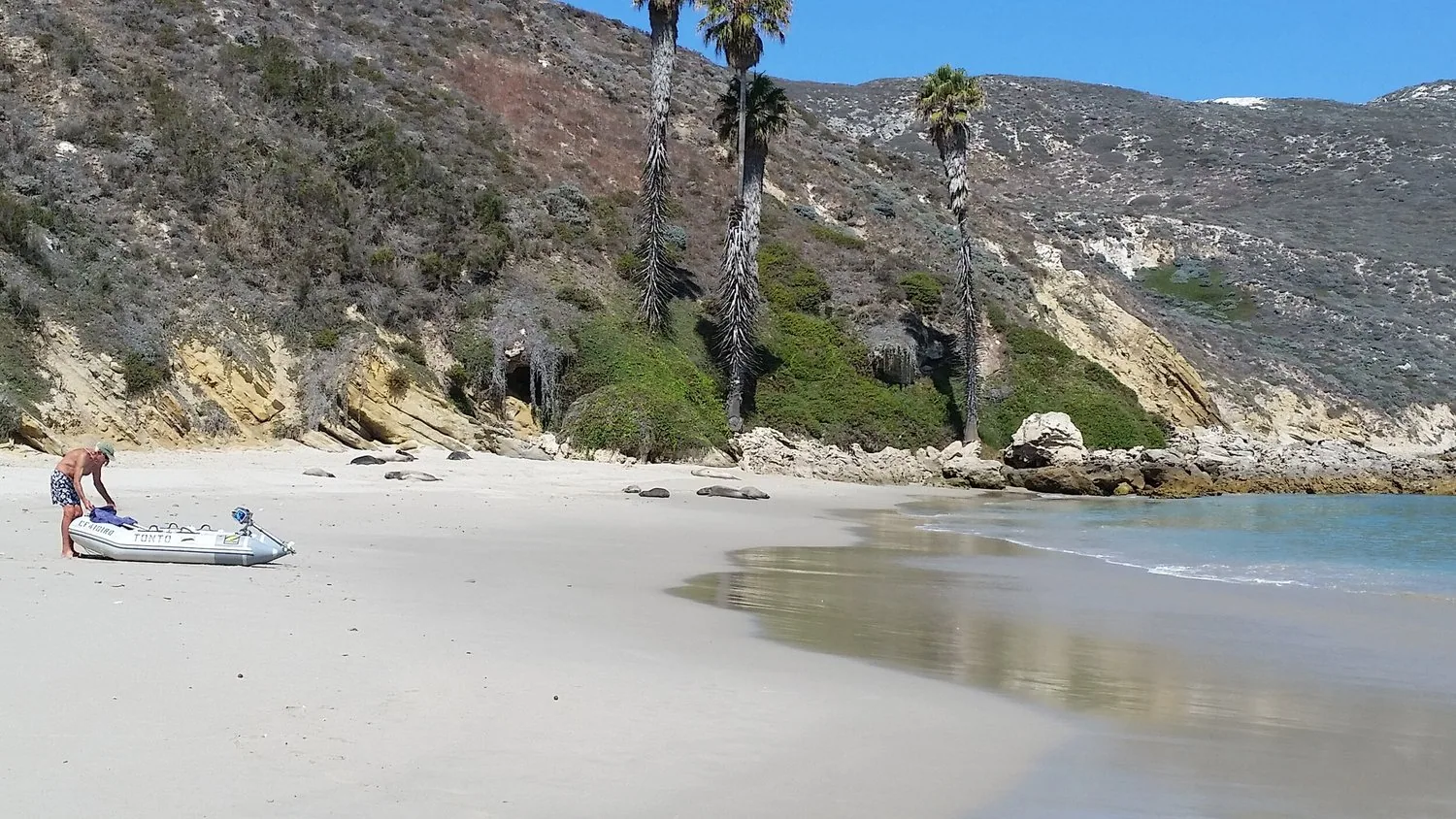 Sailor hauling an inflatable dinghy up a sandy cove beach beneath palms and cliffs at Catalina Island