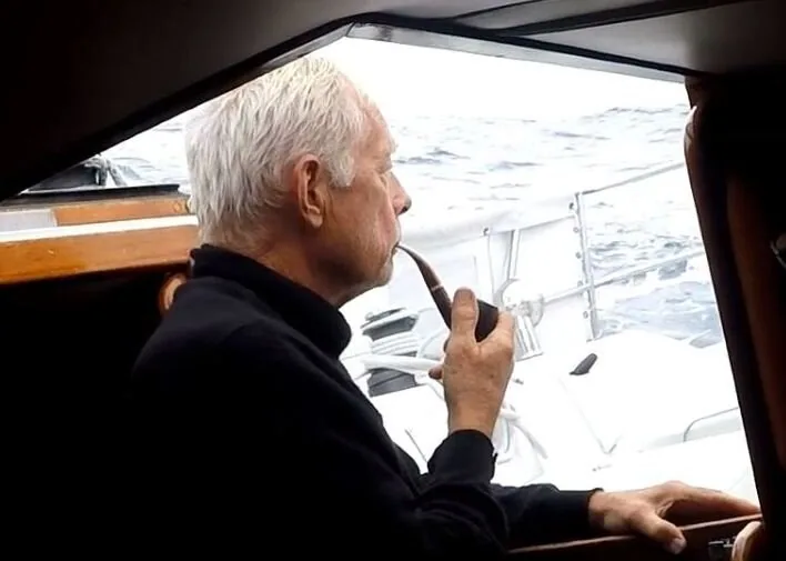 Christian Williams, white-haired and smoking a pipe, looking out to sea from the companionway