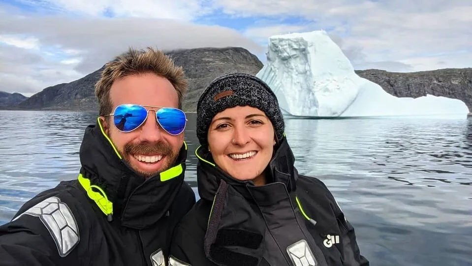 Two sailors in black foul-weather jackets take a smiling selfie in front of an iceberg in Greenland