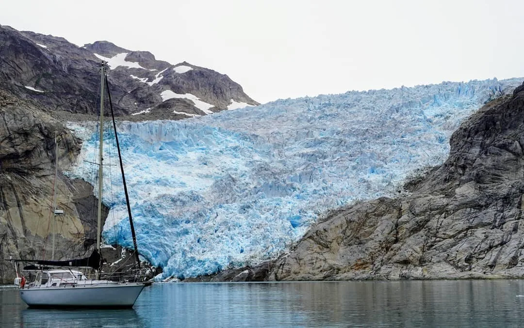 Sailboat anchored below the blue ice of a glacier tumbling between rocky slopes in Greenland