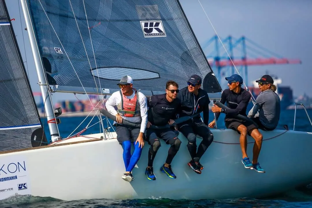 Five crew hiking on the rail of a Platu 25 under grey UK Sailmakers sails, container cranes behind