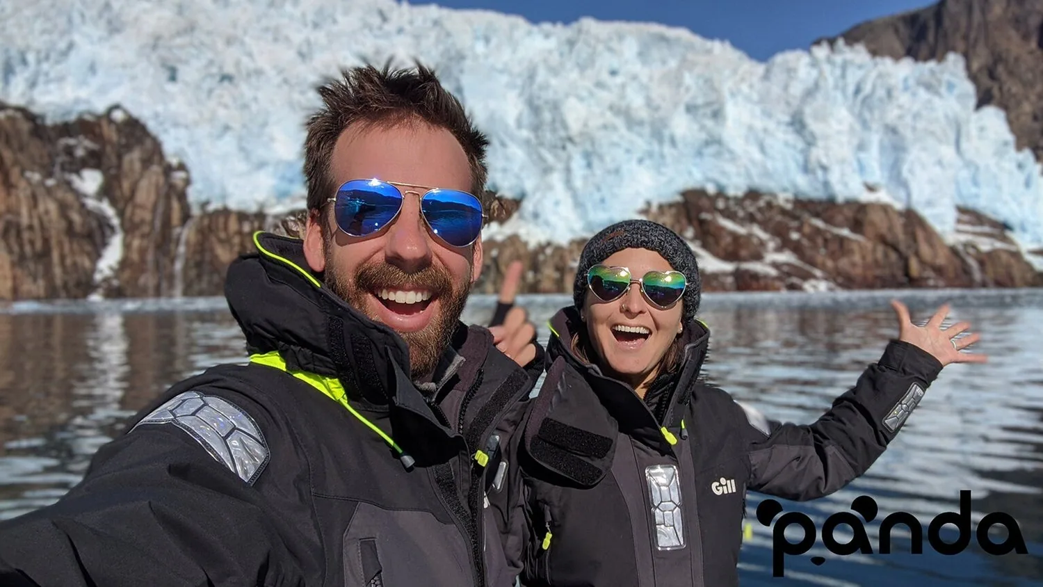 Two grinning sailors in foul-weather jackets and sunglasses pose before a glacier face, Sailing Panda logo