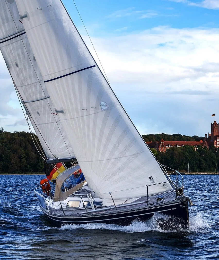 Comfortina 39 heeled under white X-Drive® Silver sails, a red-brick tower on the wooded shore behind