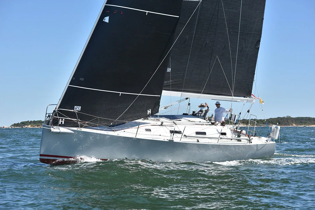 White sloop sailing upwind doublehanded under black mainsail and jib, wooded shore in the distance