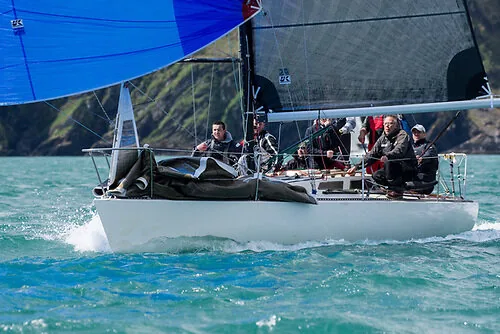 White racing sloop reaching under a blue asymmetric spinnaker and grey mainsail, full crew aboard, green hills behind