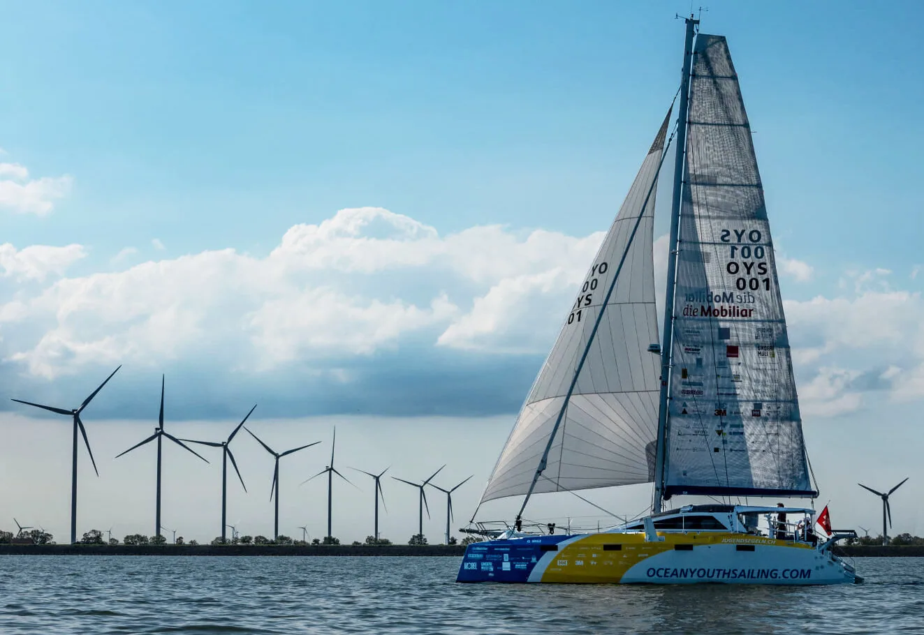 Blue-and-yellow Ocean Youth Sailing catamaran OYS 001 under grey X-Drive® mainsail and genoa, passing a row of wind turbines