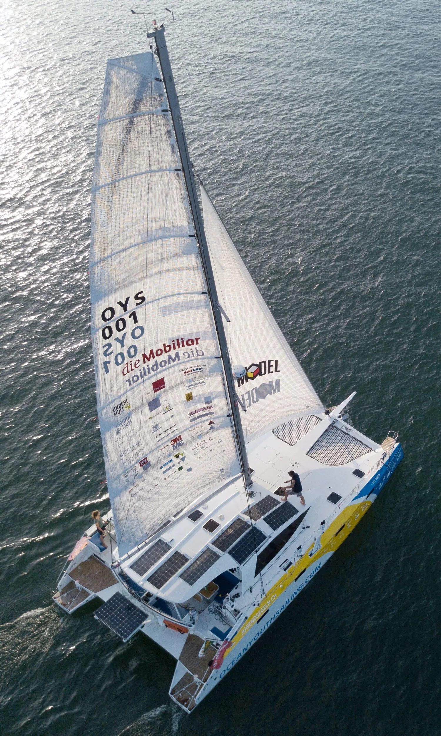 Masthead view down onto the Ocean Youth Sailing catamaran's grey X-Drive® mainsail with sponsor logos, solar panels on deck