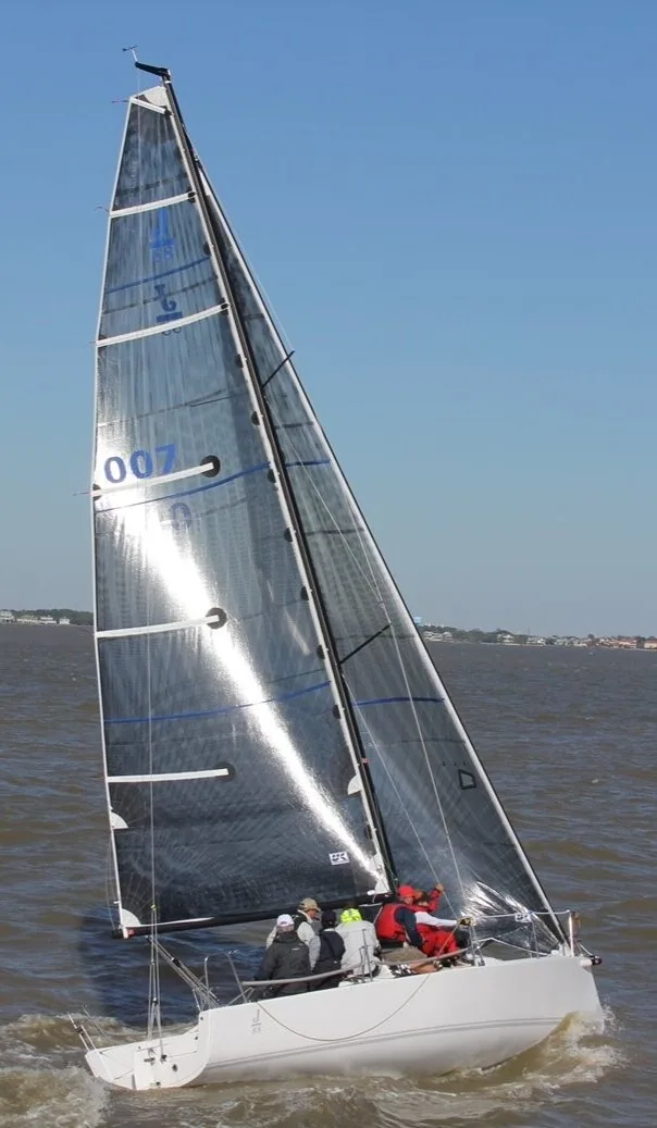 J/88 007 beating upwind under grey carbon sails, four crew in red and white in the cockpit, houses on the shore behind