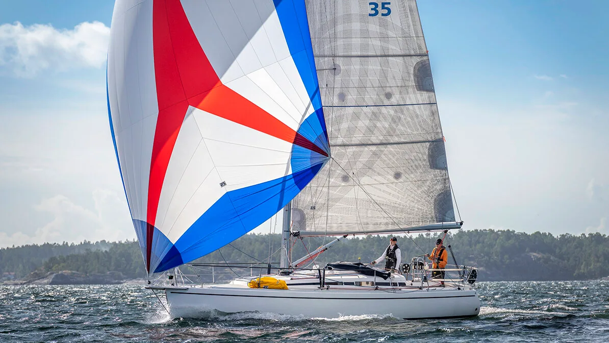 Sloop flying a red, white and blue asymmetric spinnaker with grey mainsail, two crew aboard, wooded islands behind