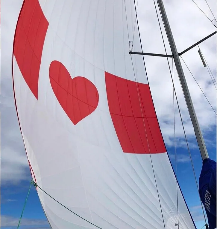 White spinnaker with a red heart between two red squares, seen from the deck against a blue sky