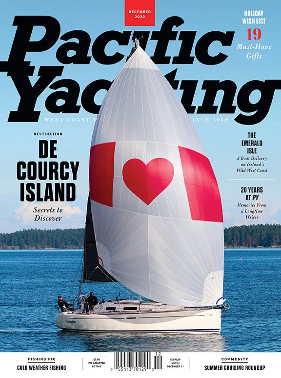 Pacific Yachting December 2020 cover: sloop flying a white spinnaker with a red heart off a wooded shore