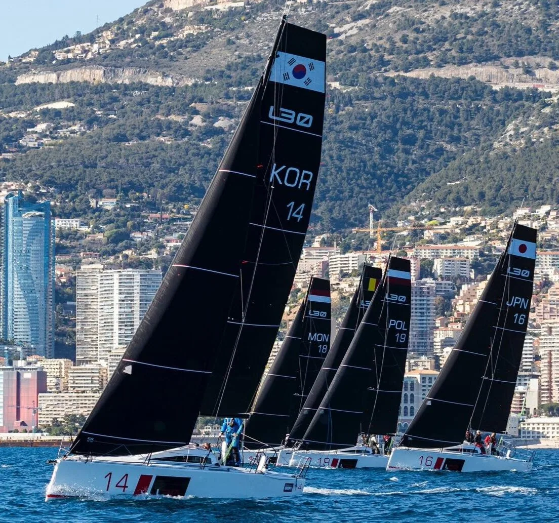 Four L30 one-designs with black sails racing upwind, KOR 14 leading, before a high-rise waterfront city backed by mountains