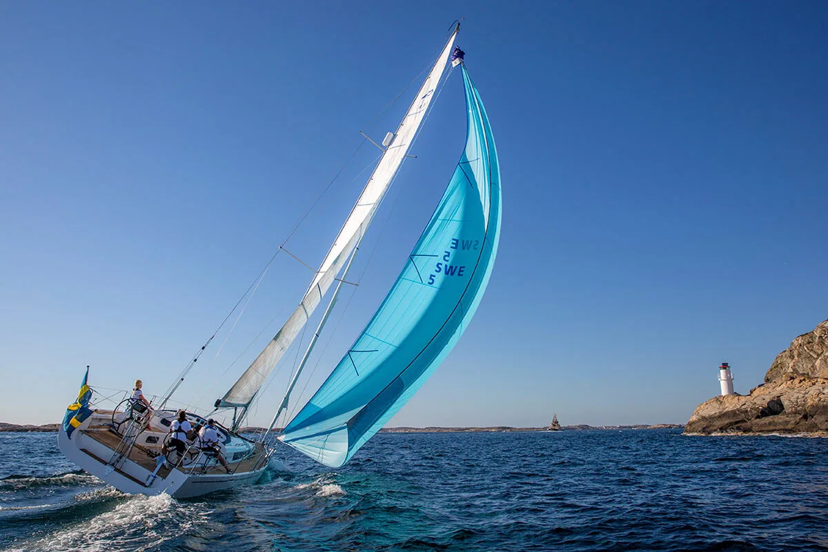 Arcona 435 reaching under a light blue asymmetric spinnaker, dousing sock bunched at the head, lighthouse on the rocks astern