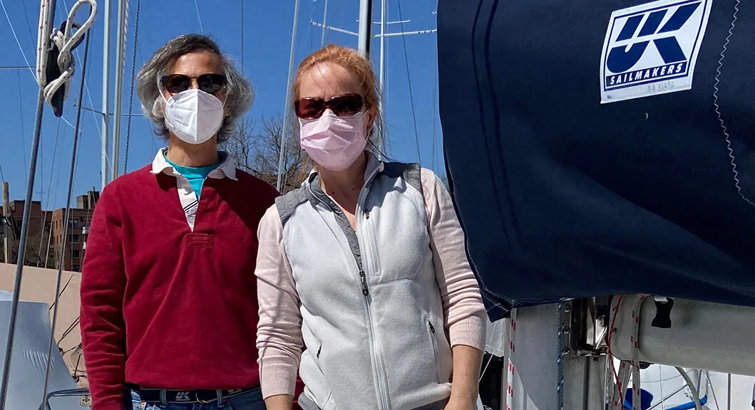 Two sailors in face masks stand on deck beside a navy sail cover bearing the UK Sailmakers logo at a marina