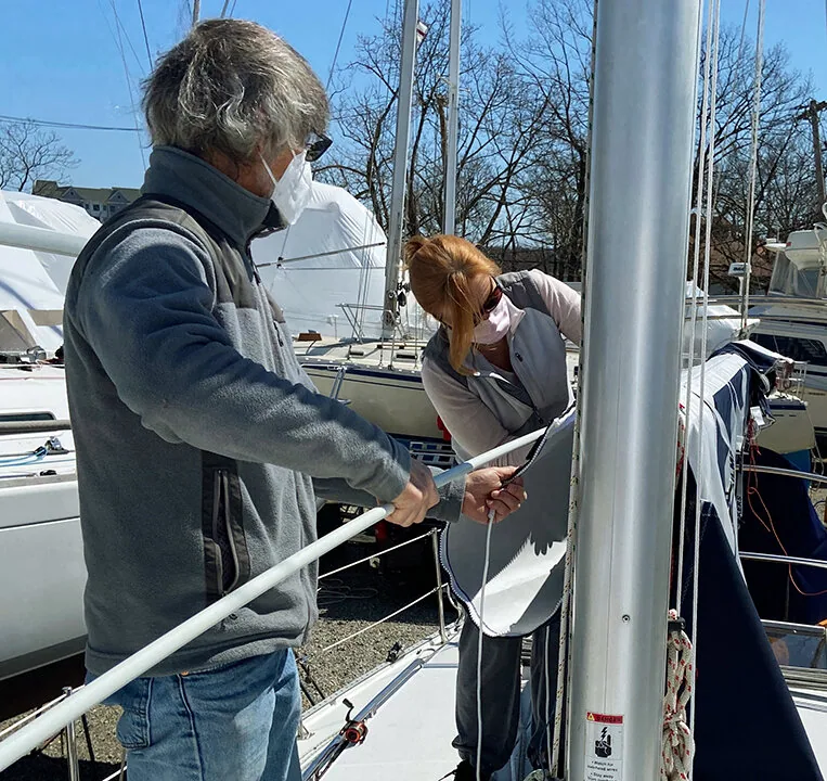 Two sailors at the mast feeding a batten into a mainsail stowed in its lazy cradle in a springtime boatyard
