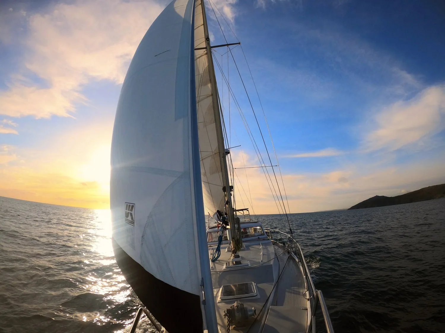 Wide-angle view down the deck of a cruising yacht under full white genoa at sunset, headland off to starboard
