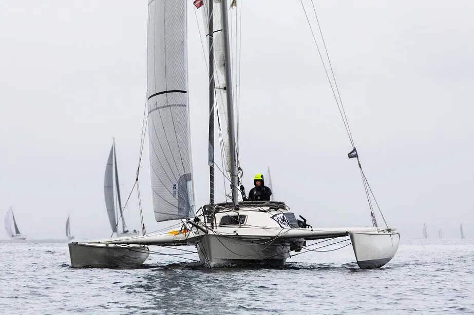 Trimaran with a lone sailor in the cockpit sailing under mainsail in grey misty conditions, fleet astern