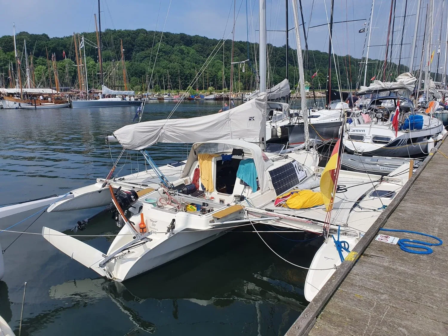 Small trimaran moored at a marina pontoon with a boom tent, German flag and solar panel, tall ships behind