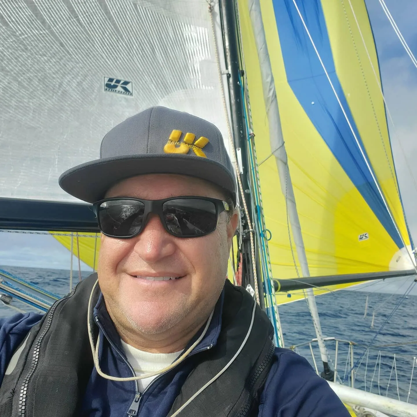 Smiling solo sailor in a UK Sailmakers cap and sunglasses, yellow and blue spinnaker and white mainsail set behind him